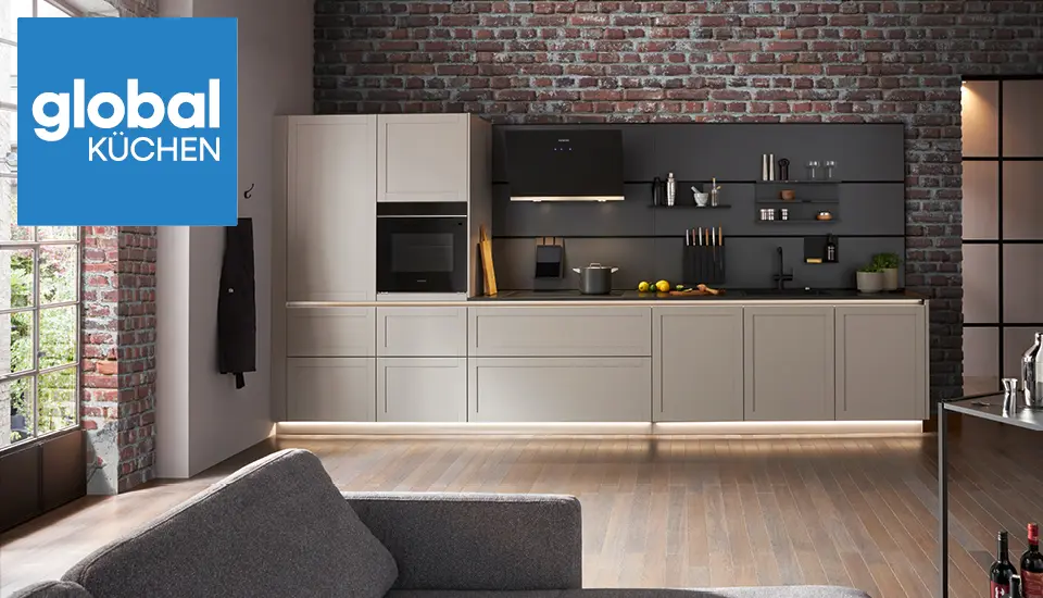 Moderne Küchenzeile mit grauen Fronten und offenen Regalen vor einer Ziegelwand, darunter LED-beleuchteter Sockel, im Vordergrund ein Sofa, Markenlogo „global KÜCHEN“ oben links.